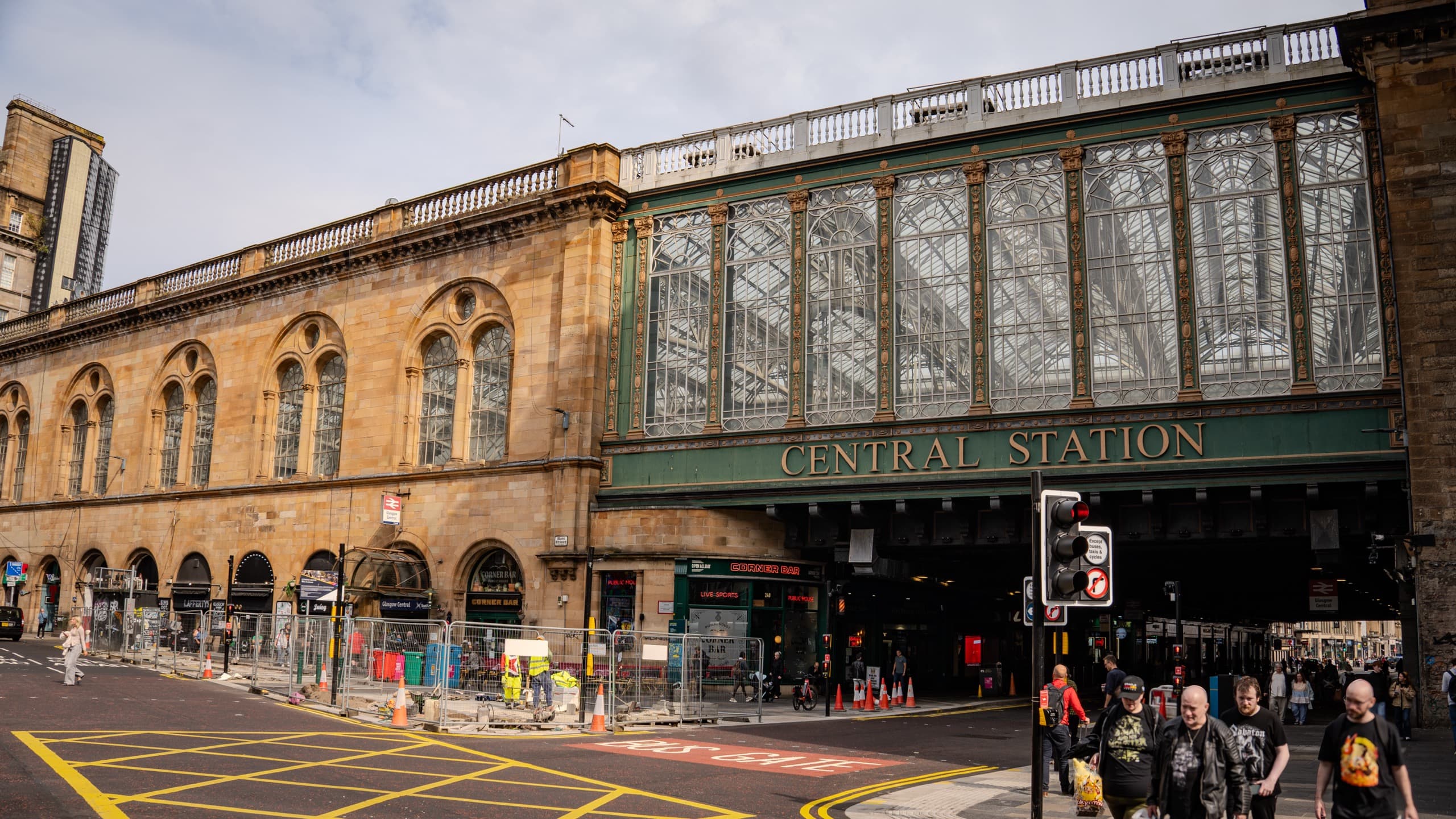 Central Station in Glasgow, Scotland. An older brick building with ornate arching windows and several storefronts built into the side. The building extends over a street where signage says "Central Station". The overhang area above the signage is almost entirely glass.