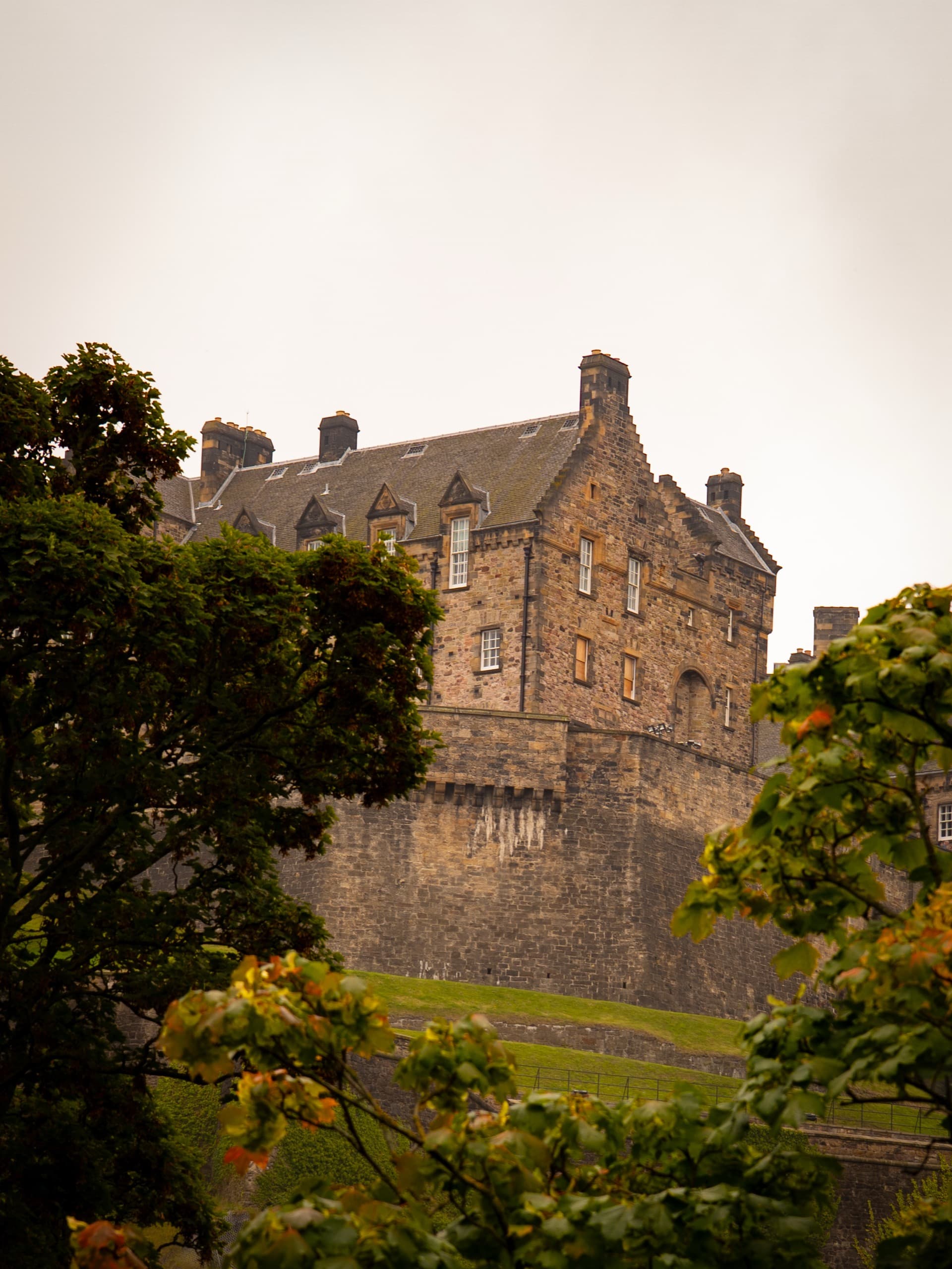 A corner of a brick and stone castle perched up on a hill with a wall around it. There are trees in the foreground.