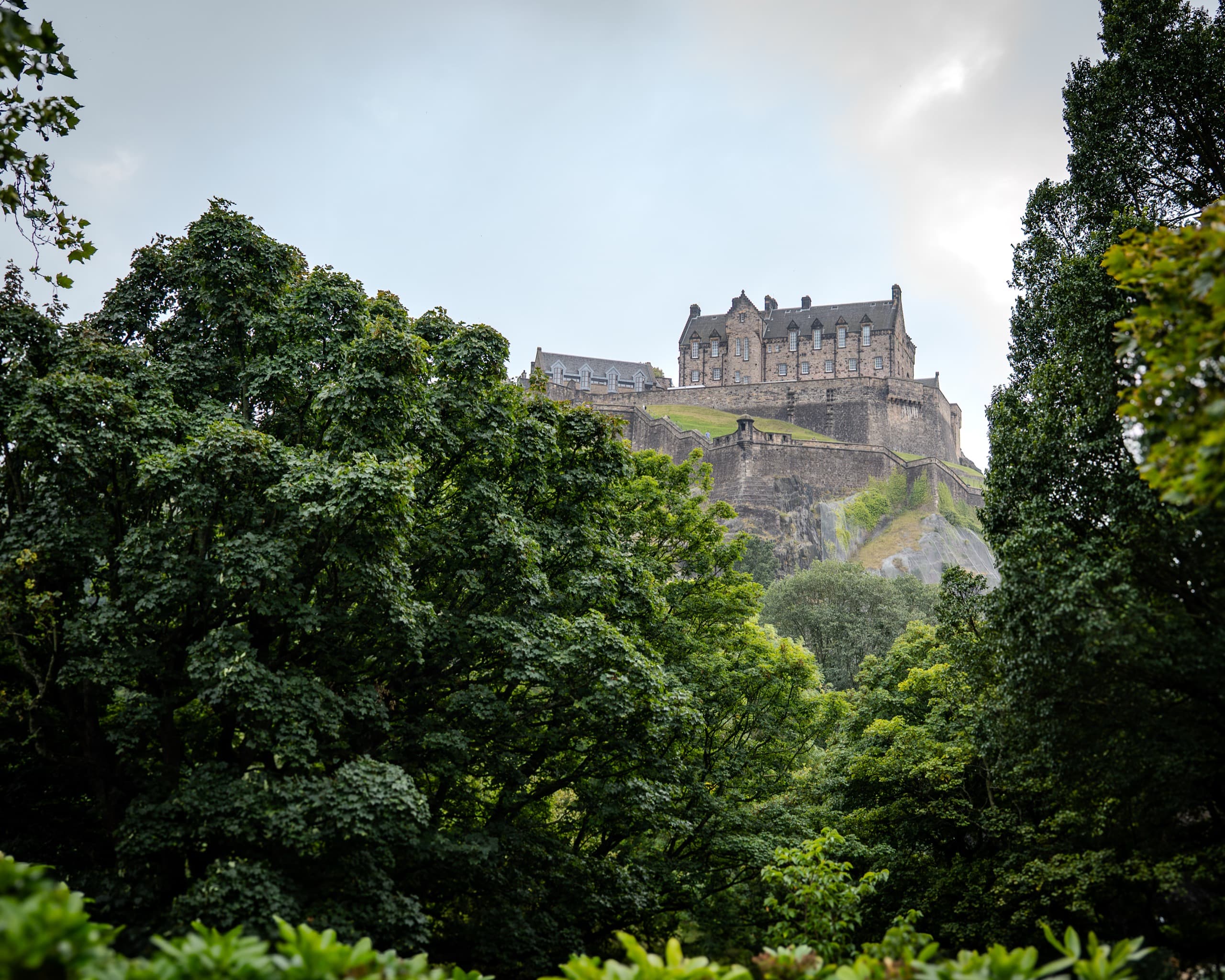 A brick and stone castle sitting on top of a hill on a cloudy day. There are walls immediately surrounding the castle and trees after that.