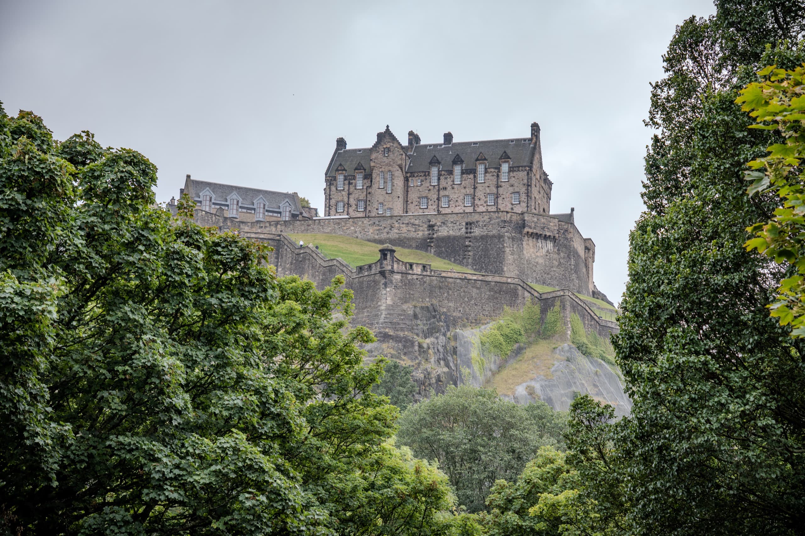 A brick and stone castle sitting on top of a hill on a cloudy day. There are walls immediately surrounding the castle and trees after that. The castle is more prominent in frame than the previous photo.