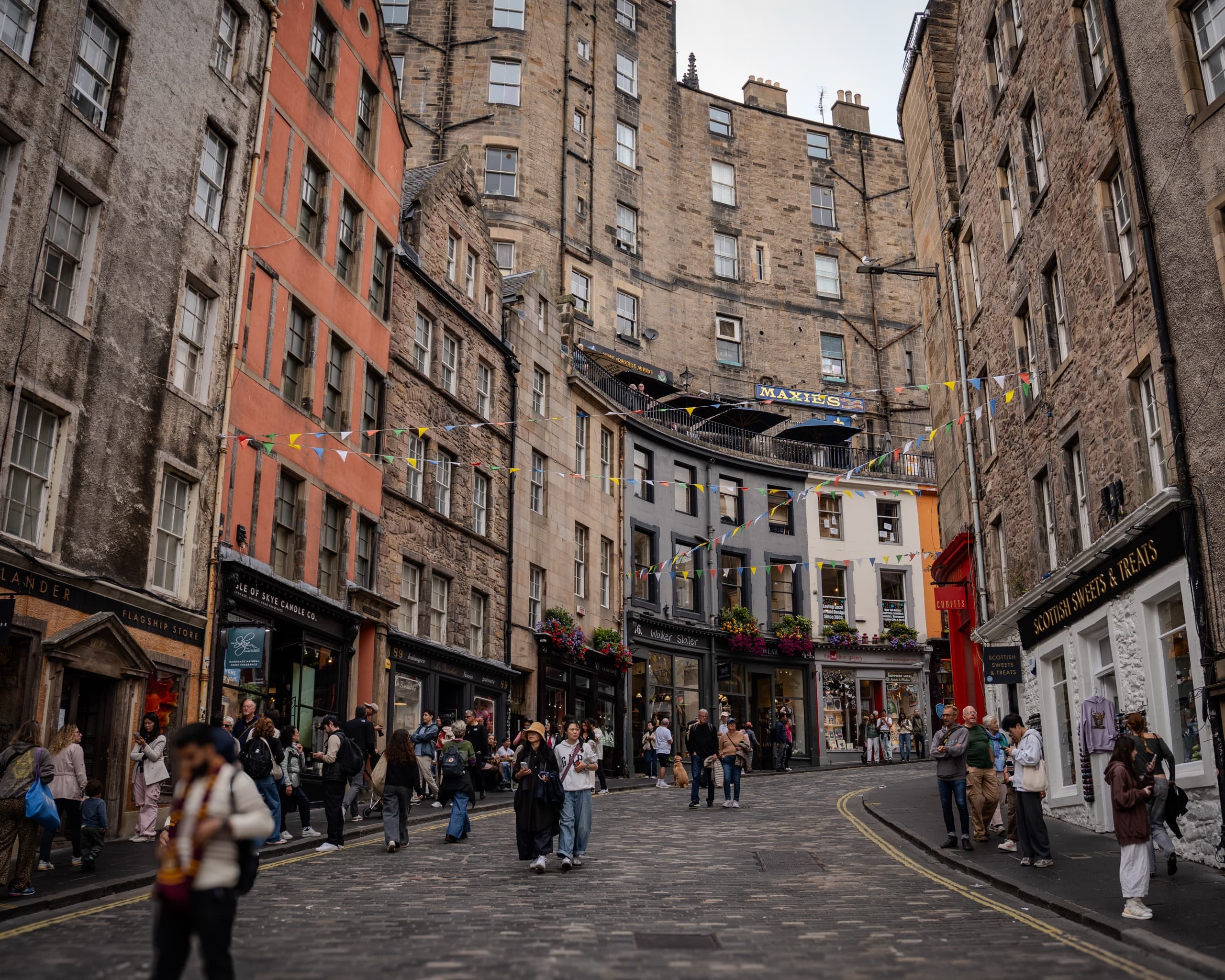 Cobblestone street in Edinburgh, Scotland. There are old stone buildings along both sides as the street ascends into a hill and turns. Many people are walking along the street and there are several storefronts along the sides.