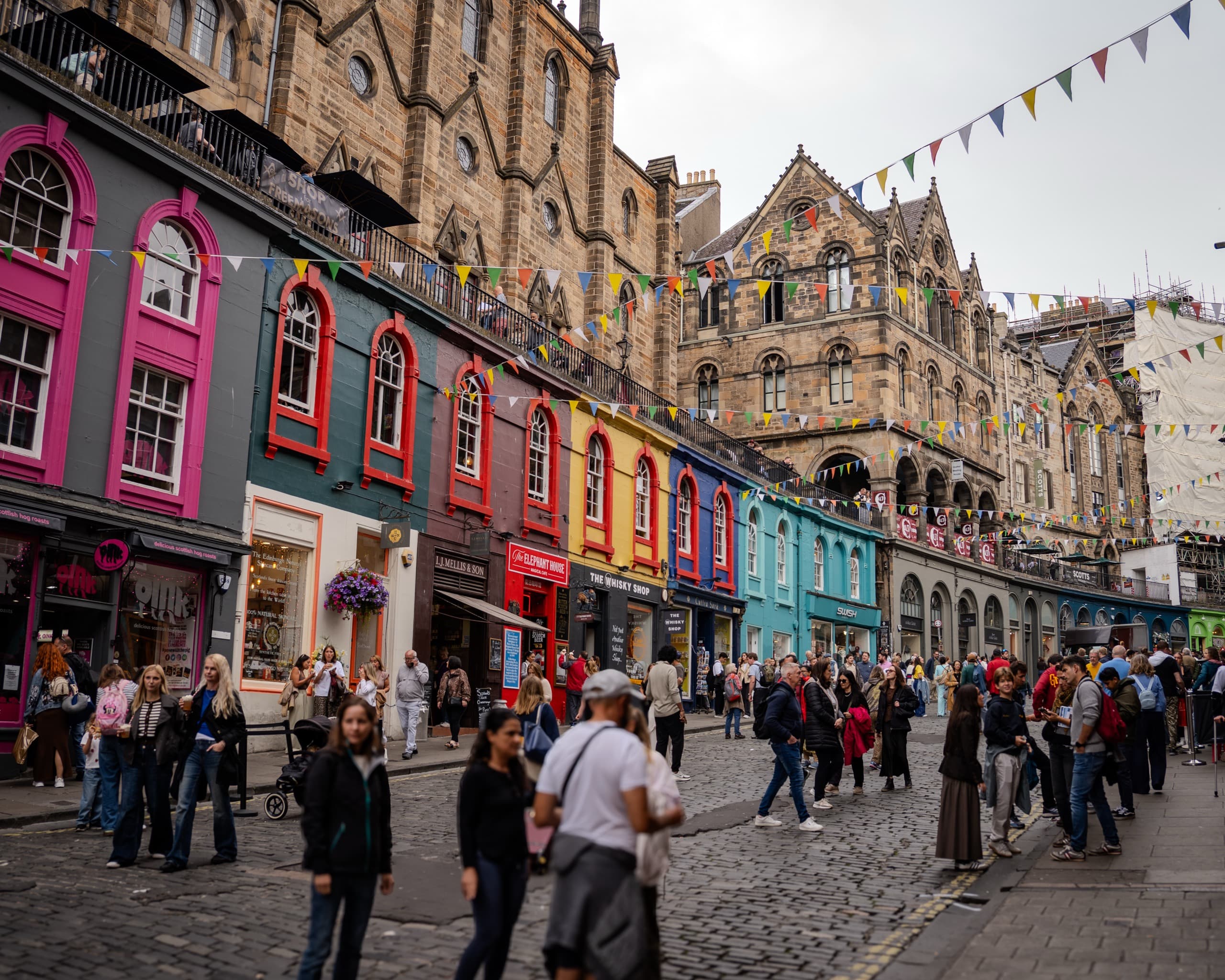 A stone-paved street in Edinburgh with brightly painted buildings bunched up next to each other. There are many people out on the street.