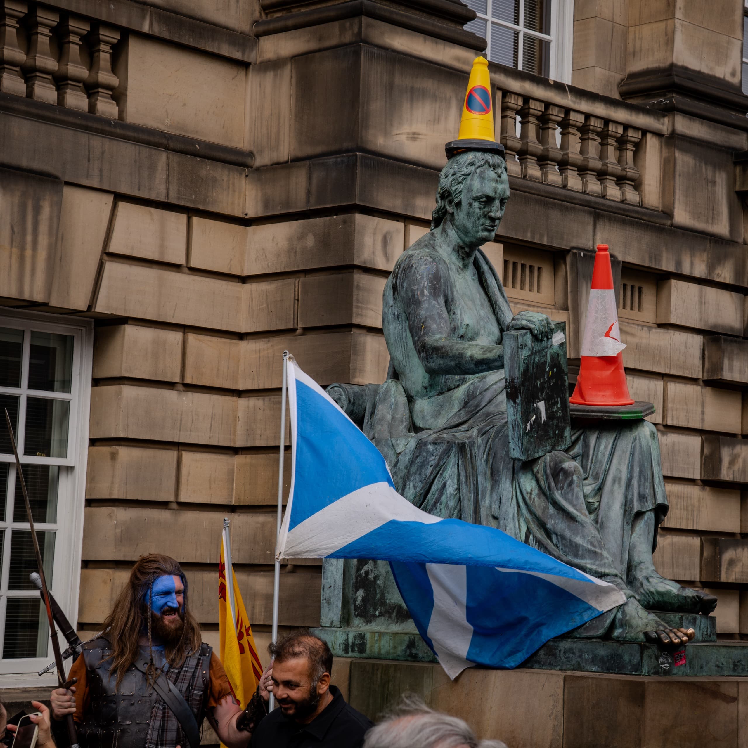 A man in traditional Scottish attire, including the blue half-face paint, sword, and kilt, next to a statue of a man. Of course, there are traffic cones on the statue. The Scottish flag is in front of them.