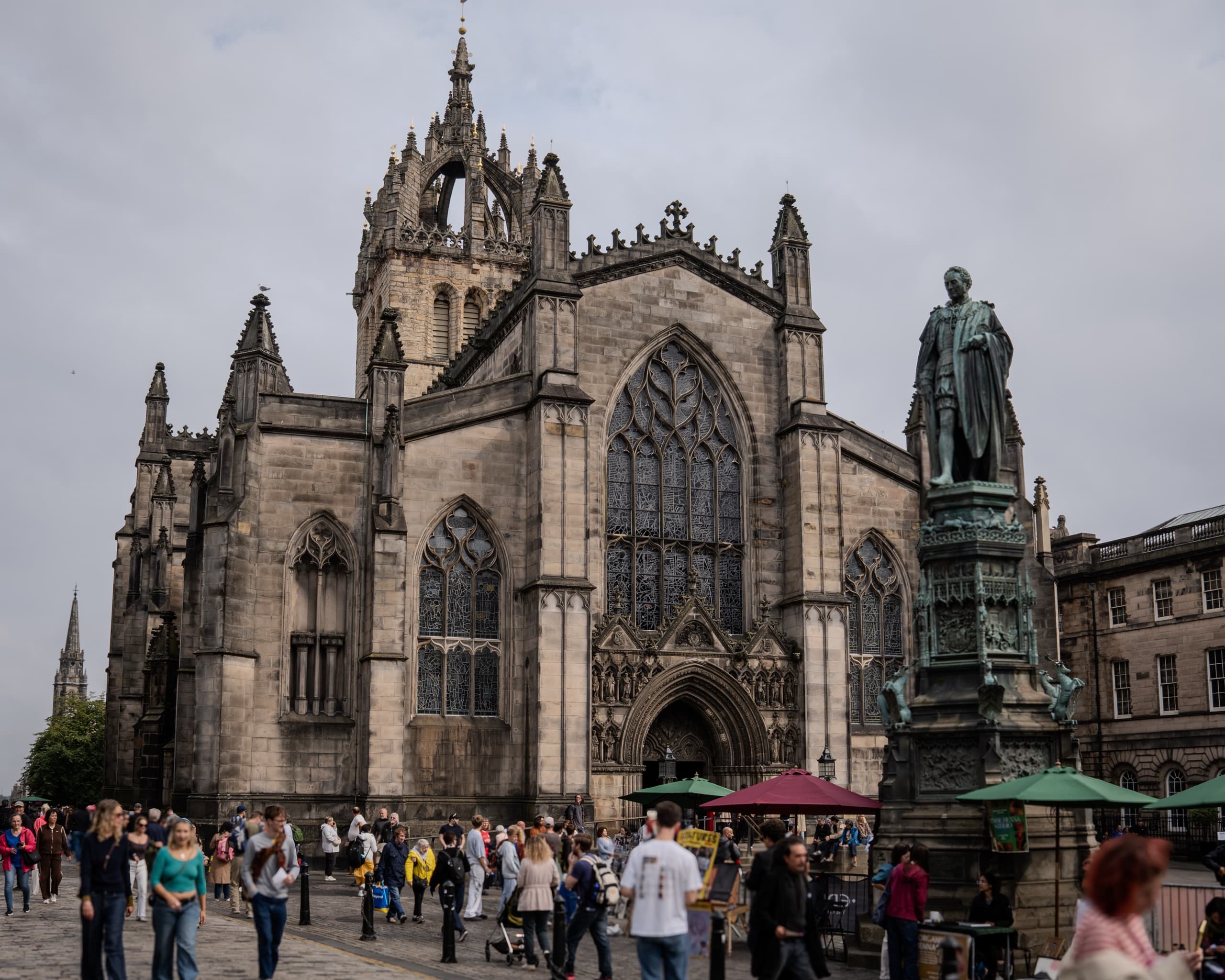 A massive stone cathedral in a courtyard of Old Town Edinburgh. There is a statue of a man in front and a large crowd walking around the courtyard.