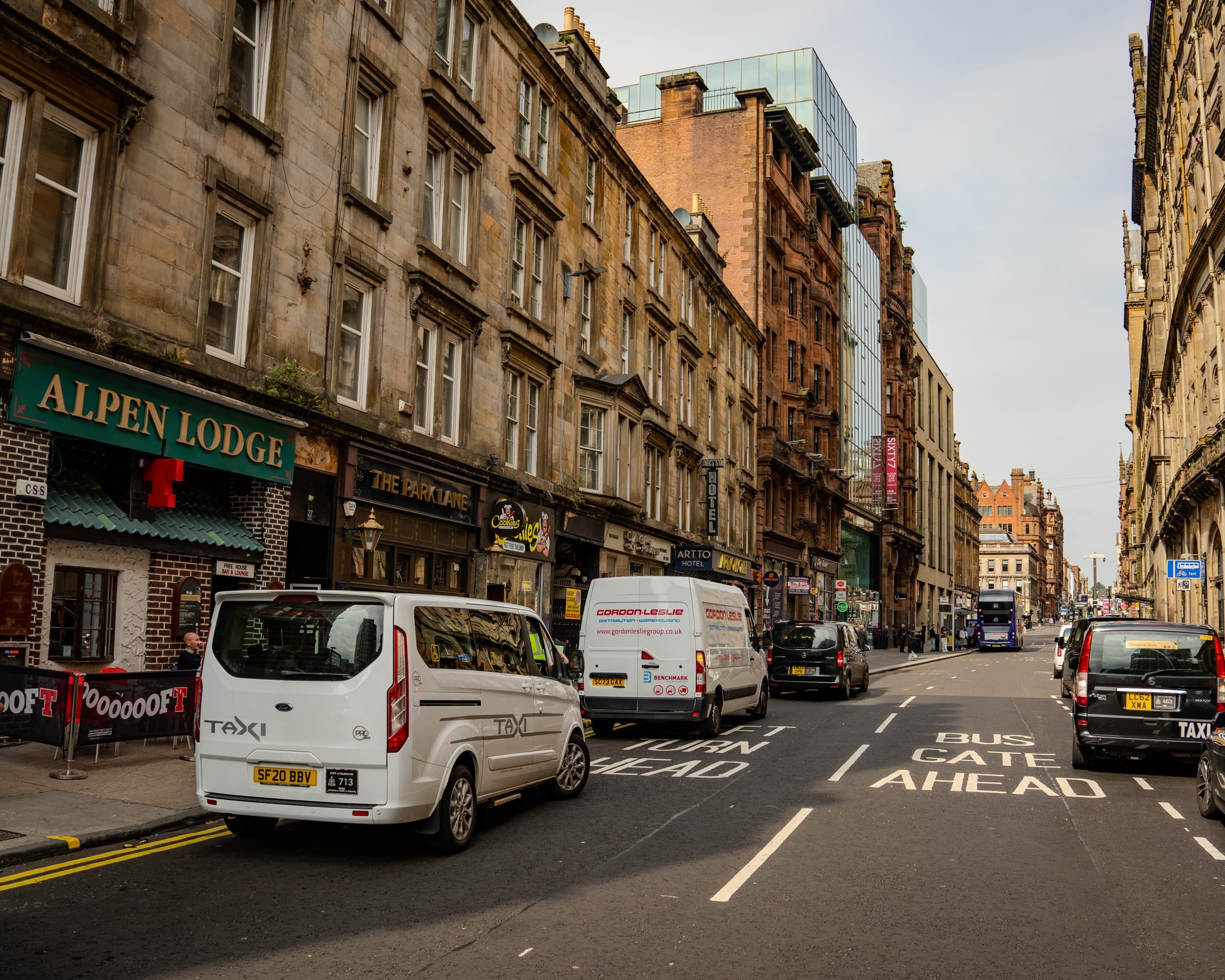 Looking down a street in Glasgow, Scotland. There are several vans parked along the side of the street, servicing storefronts and bars.
