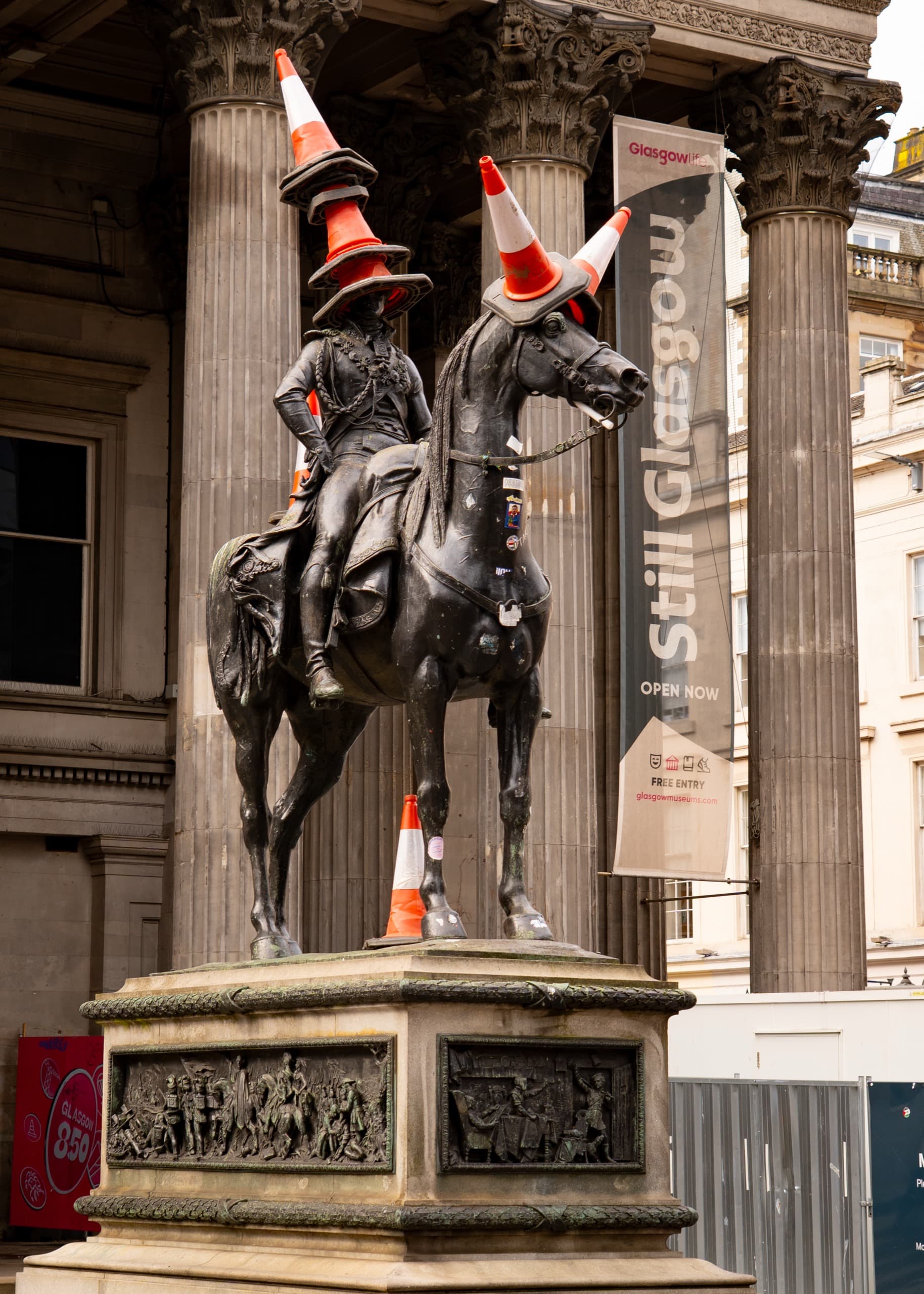 Statue of a man in military regalia on a horse in front of the Gallery of Modern Art in Glasgow. This statue is covered in traffic cones. Any spot a traffic cone can fit, there is one.
